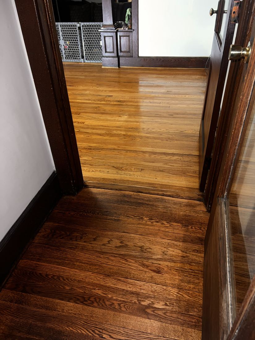 Finished stained hardwood hallway leading into a finished natural hardwood room